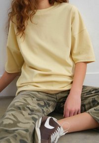 Camiseta de algodón amarilla de gran tamaño con mangas cortas, combinada con pantalones de camuflaje verde y zapatillas marrones con detalles blancos.