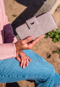 Žena v růžovém svetru a modrých džínách drží prošívanou peněženku v barvě mauve s černým logem a sedí venku v slunečném dni.