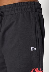 Calças de moletom pretas feitas de tecido jersey, com um cós elástico, bolsos laterais e um pequeno logótipo branco na perna. Acento em texto vermelho.