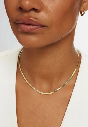 Halskette aus goldfarbenem Metall mit glattem, flachem Band und leicht gebogener Form, getragen auf einer Haut mit neutralem Ton und weißer Kleidung.