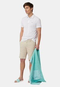 Polo bianco, pantaloni corti beige e sandali a righe. Tiene un asciugamano turchese con dettaglio a tasca. Tessuto morbido e vestibilità rilassata.