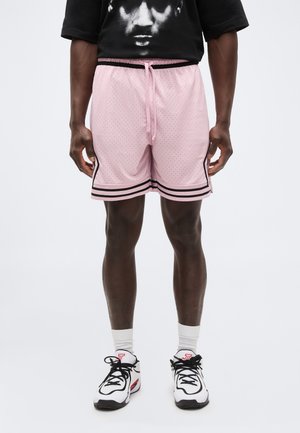 Άνδρας που φοράει ανοιχτό ροζ σορτς μπάσκετ με μαύρες ρίγες, λευκές κάλτσες, λευκά και μαύρα αθλητικά παπούτσια και μαύρο κοντομάνικο μπλουζάκι με σχέδιο.