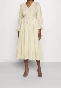 Abito midi a righe giallo chiaro con scollatura a V, maniche lunghe, polsini arricciati e vita annodata. Presenta una gonna a strati e una vestibilità comoda.