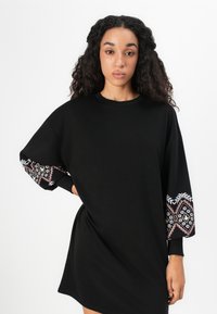 Zwart oversized sweatshirt jurk met decoratieve patroon mouwen die witte en roze ontwerpen bevatten. Zachte stof, relaxed fit en ronde halslijn.