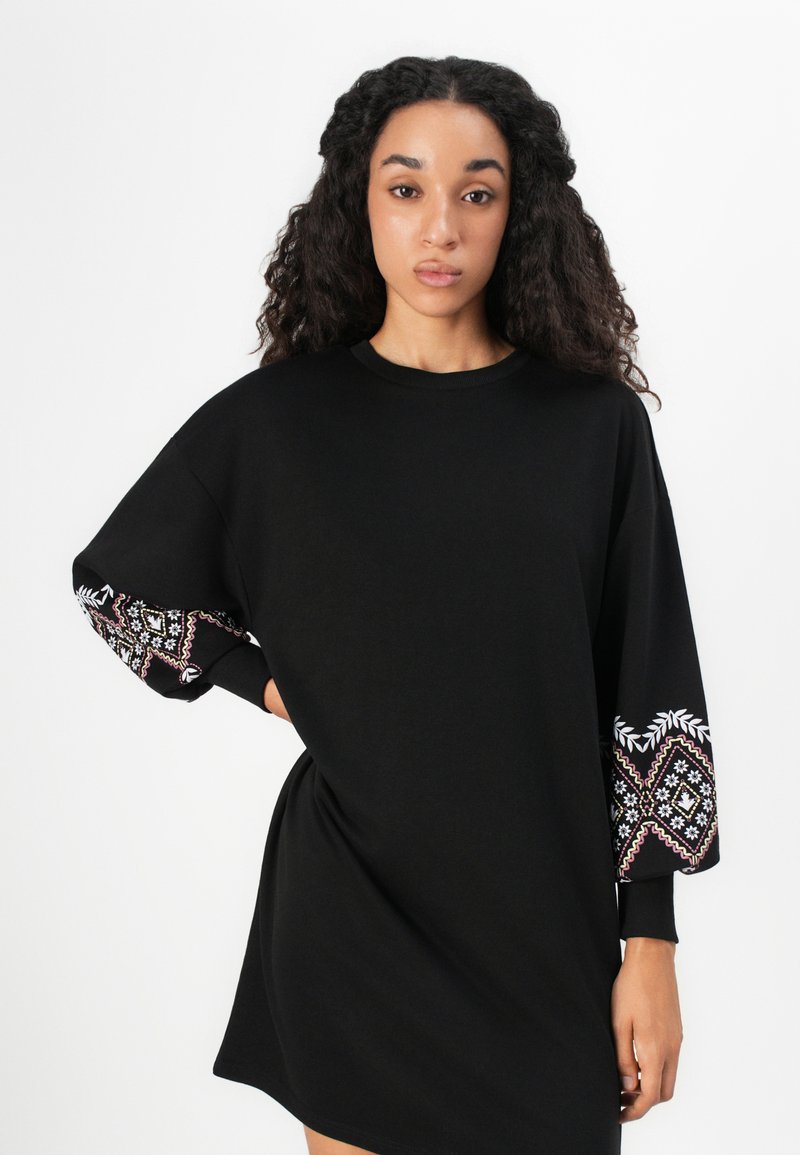 Abito felpa nero oversize con maniche decorative a motivi che presentano disegni bianchi e rosa. Tessuto morbido, vestibilità comoda e scollo tondo.