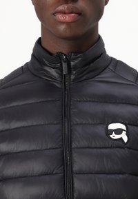 Gepolsterte schwarze Jacke mit hohem Kragen und einem Frontreißverschluss. Zeigt ein kleines weißes Logo-Patch mit einer Figur in Sonnenbrille.