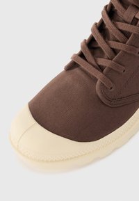 Zapatillas de lona marrón con una puntera redonda de goma crema, cordones texturizados y una suela cosida. Vista en primer plano que resalta el material y el diseño.