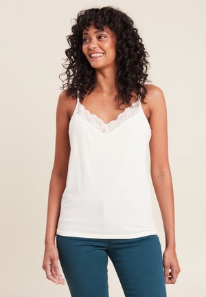 Femme souriante aux cheveux bouclés portant un haut sans manches blanc à bordure en dentelle et un pantalon bleu sarcelle, debout devant un fond uni.