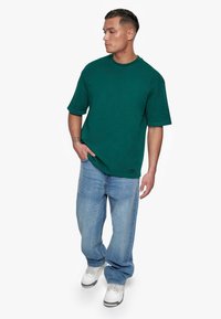 Grøn kortærmet t-shirt i en afslappet pasform, kombineret med lyseblå jeans og hvide sneakers. T-shirten har et lille logo-label.