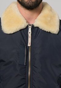 Bomber en marine avec un col en fourrure crème, une fermeture éclair dorée et une poche à rabat. Texture lisse avec une finition légèrement brillante.