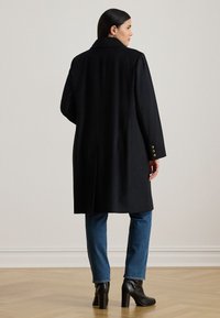 Long black coat with a classic collar, three gold button accents on the sleeves, and a straight cut, paired with blue jeans and black heeled boots.