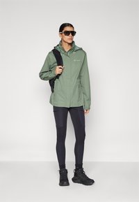 Lys grønn vanntett jakke med frontglidelås, hette og glidelåslommer på sidene, kombinert med svarte leggins og solide svarte turstøvler.