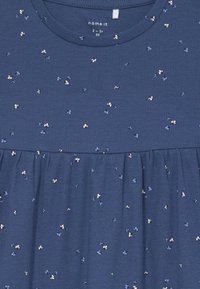 Rövid ujjú sötétkék felső, bordás textúrával, ráncolt derékkal és világoskék, valamint fehér virágmintával. Márkacímke látható.