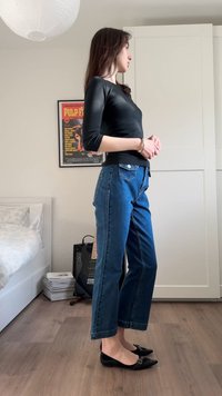 Γυναίκα που στέκεται σε δωμάτιο φορώντας μαύρη μπλούζα με μακριά μανίκια, μπλε τζιν και μαύρα φλατ παπούτσια, δίπλα σε ένα κρεβάτι και έναν ενισχυτή κιθάρας.