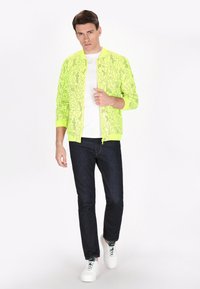 Jachetă bomber galbenă neon cu un model geometric, purtată cu un tricou alb, blugi închiși și adidași albi.