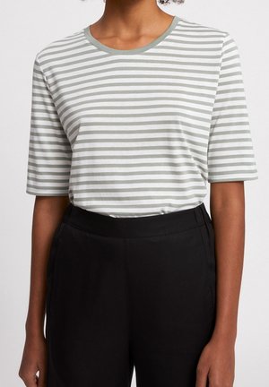 Personne portant une chemise à manches courtes, blanche et grise claire à rayures horizontales, rentrée dans un pantalon noir taille haute.