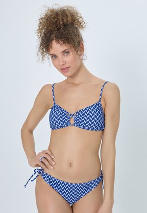 Young woman with curly hair wearing a blue and white patterned bikini with side ties, posing against a plain light background.
