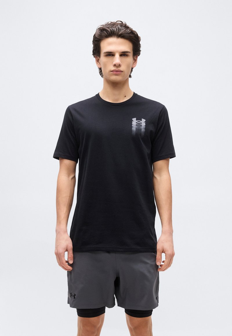 Νεαρός άνδρας όρθιος, φορώντας μαύρο μπλουζάκι Under Armour και σκούρα γκρι σορτς με μαύρα συμπιεστικά κολάν από κάτω.