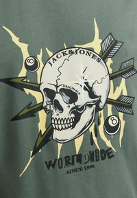 T-shirt graphique vert avec un design de crâne comprenant des flèches, des éclairs et du texte. L'impression inclut des accents blancs, noirs et jaunes.