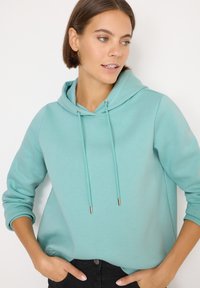 Felpa verde menta con cordini regolabili e dettagli in metallo argentato, caratterizzata da una vestibilità rilassata e polsini a coste, abbinata a pantaloni neri.