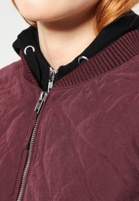 Giacca bomber borgogna trapuntata con zip argentata, collo a costine e felpa nera sotto, caratterizzata da tessuto testurizzato e motivo a diamante.