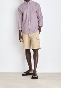 Camicia a maniche lunghe a quadretti rossi e bianchi, pantaloni cargo beige con tasche e sandali neri a infradito, in piedi su un pavimento neutro.