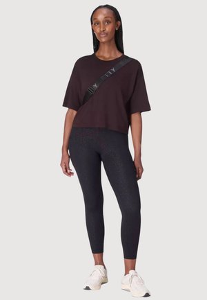 Jonge vrouw staand, draagt een los donker T-shirt, zwarte legging met patroon, witte sneakers en een zwarte crossbodyriem op een neutrale achtergrond.