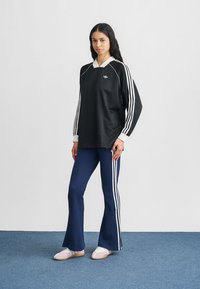 Sweatshirt preta oversize com riscas brancas e logótipo, combinada com calças azuis caneladas boca de sino com riscas laterais, e sapatos brancos.