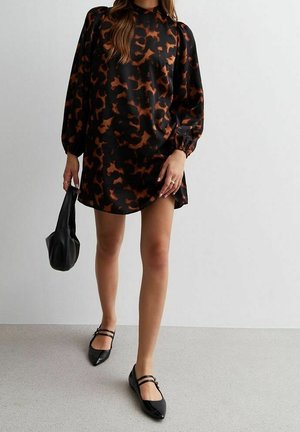 Frau trägt ein langärmeliges Kleid mit schwarzem und braunem Muster, hält eine schwarze Handtasche und trägt schwarze Mary Jane Ballerinas.