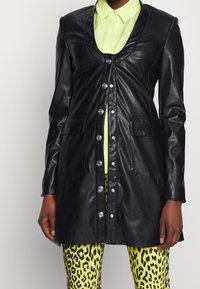 Cappotto lungo in pelle nera con scollo a barca, bottoni frontali, due tasche e una texture liscia, indossato sopra un top verde brillante e pantaloni con stampa leopardata.