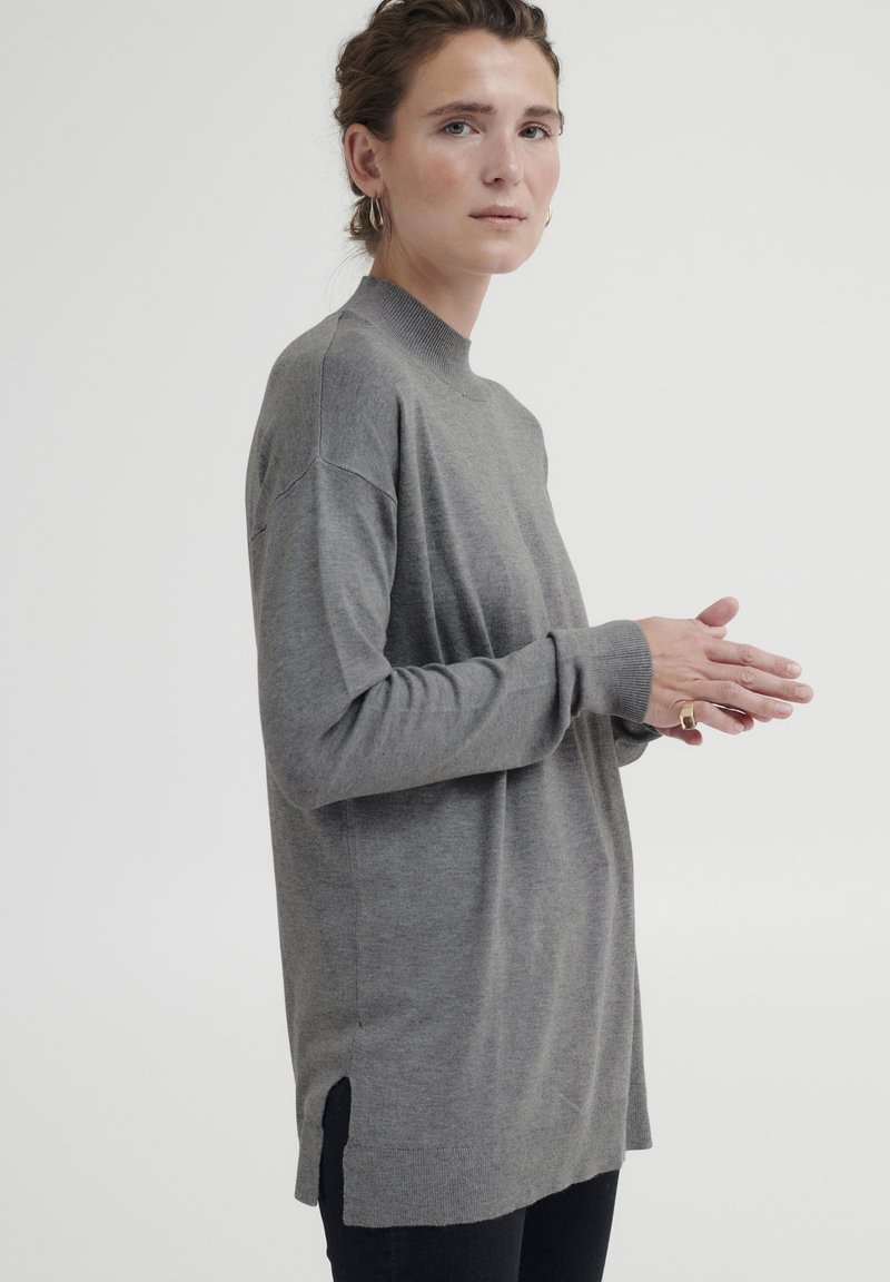 Grauer langärmeliger Pullover aus weichem Material mit hohem Kragen, seitlichen Schlitzen und gerippten Bündchen. Modell trägt dunkle Jeans.