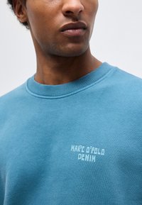 Man draagt een lichtblauwe sweatshirt met ronde hals en witte tekst "Marc O'Polo Denim" op de borst, tegen een effen lichte achtergrond.