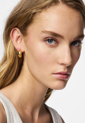 Guldtonade ringörhängen med en slät, polerad yta, buren av en modell med långt, ljust hår och ljus hy. Bakgrunden är enfärgad vit.