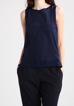 Vrouw draagt een mouwloze marineblauwe blouse en losse zwarte broek met handen in de zakken tegen een effen achtergrond.