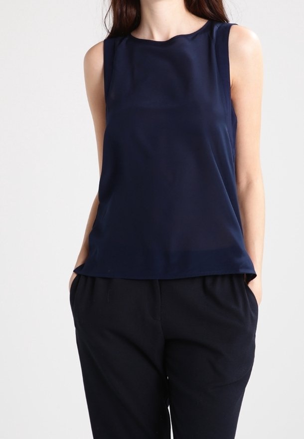Femme portant un chemisier bleu marine sans manches et un pantalon noir ample, les mains dans les poches sur un fond uni.