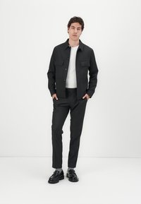 Schwarze, knöpfbare Jacke mit zwei Brusttaschen, kombiniert mit schwarzen, eng anliegenden Hosen. Das Modell trägt ein weißes T-Shirt und schwarze Schuhe.