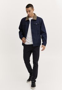 Donkerdenim jack met een bruine faux fur kraag, voorzien van knoopsluitingen en twee borstzakken; gedragen over een wit t-shirt en zwarte broek.