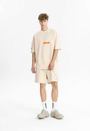 Joven con camiseta oversize beige con logo naranja, pantalones cortos a juego con detalle de cadena, calcetines blancos con texto negro y zapatos beige texturizados.