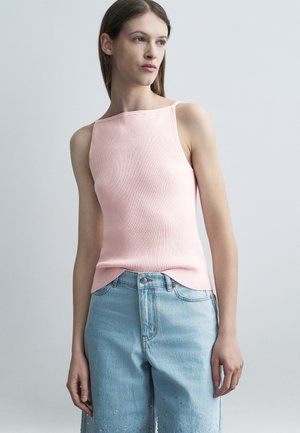 Young woman with straight brown hair wears a light pink sleeveless ribbed top and high-waisted light blue jeans with embellishments.