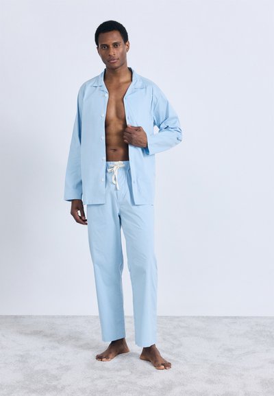 Svetlo modra, dvodelna garnitura za sproščanje, ki vključuje srajco na gumbe in hlače z elastiko in vrvico za vezanje, izdelana iz gladkega blaga.