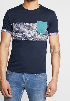 Mężczyzna ubrany w granatowy T-shirt z białym wzorem liści na klatce piersiowej, turkusową kieszonką na piersi i podwiniętymi rękawami, w połączeniu z niebieskimi dżinsami.