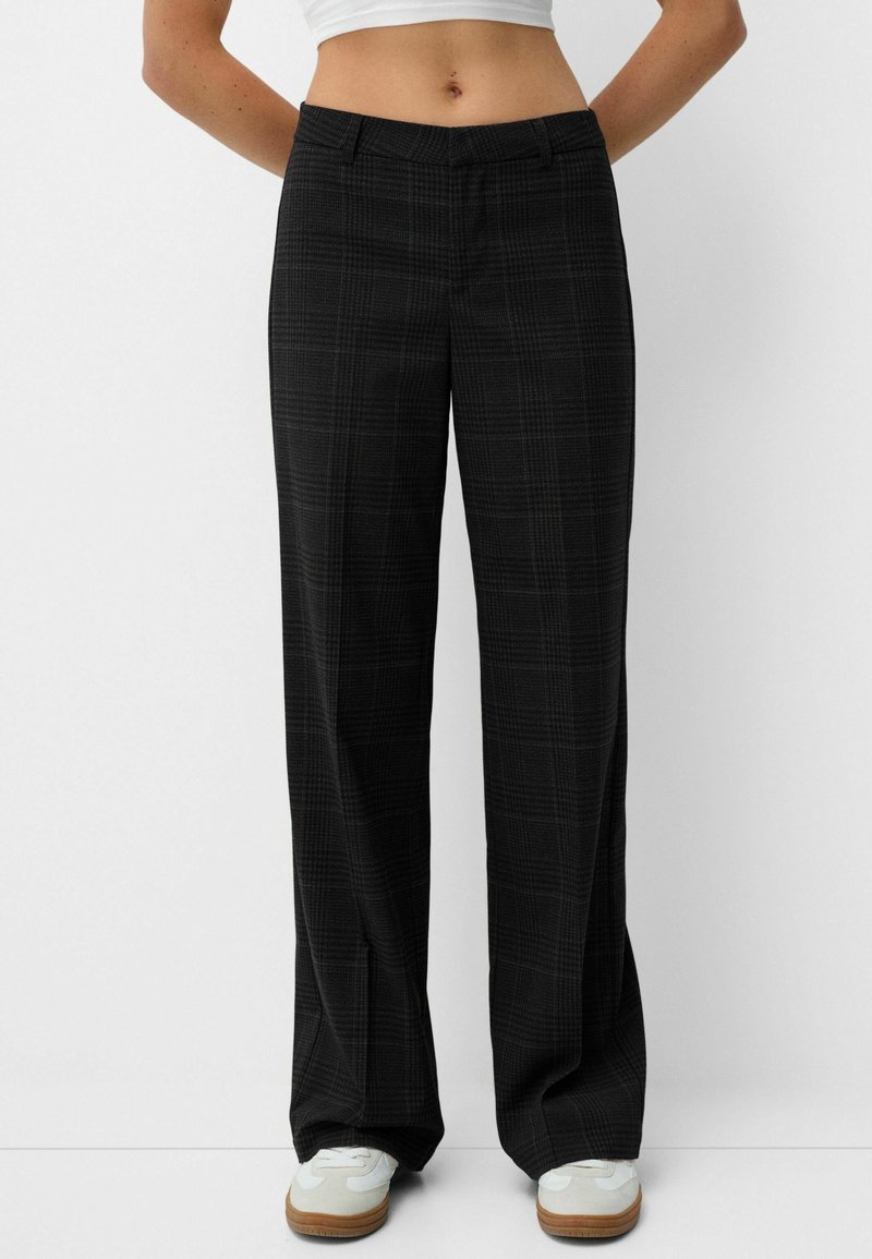 Femme portant un pantalon large à carreaux noir et des baskets blanches sur un fond uni.