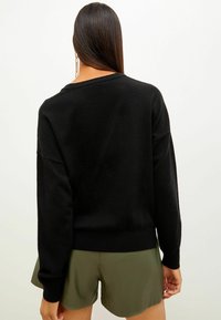 Suéter negro de gran tamaño con un tejido texturizado, mangas anchas y un hombro ligeramente caído. Combinado con pantalones cortos verdes.