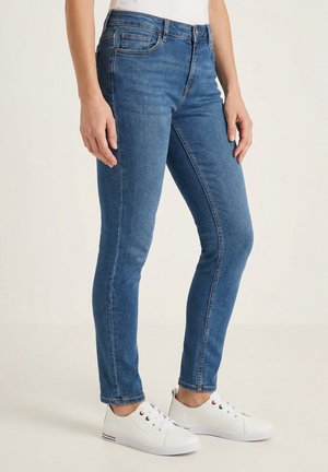 Slankzittende blauwe spijkerbroek gedragen door een persoon die staat, gecombineerd met witte lage sneakers en een wit shirt.