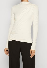 Långärmad, krämfärgad topp med hög hals och diagonalt drapering över framsidan, med en mjuk, elastisk tyg och smal passform.
