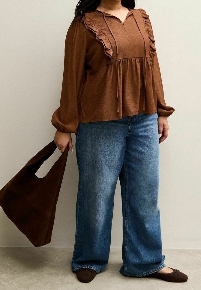 Personne portant un chemisier marron à manches longues avec volants, un jean bleu à jambes larges, des ballerines marron, et tenant un grand sac fourre-tout marron en daim.