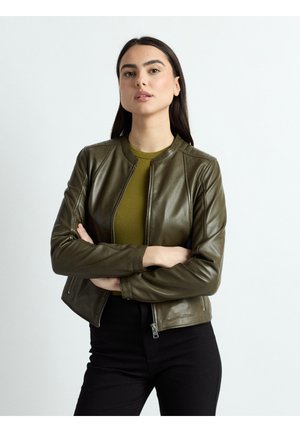 Vrouw met lang donker haar, gekleed in een olijfgroene leren jas en bijpassende trui, staand met gekruiste armen tegen een witte achtergrond.