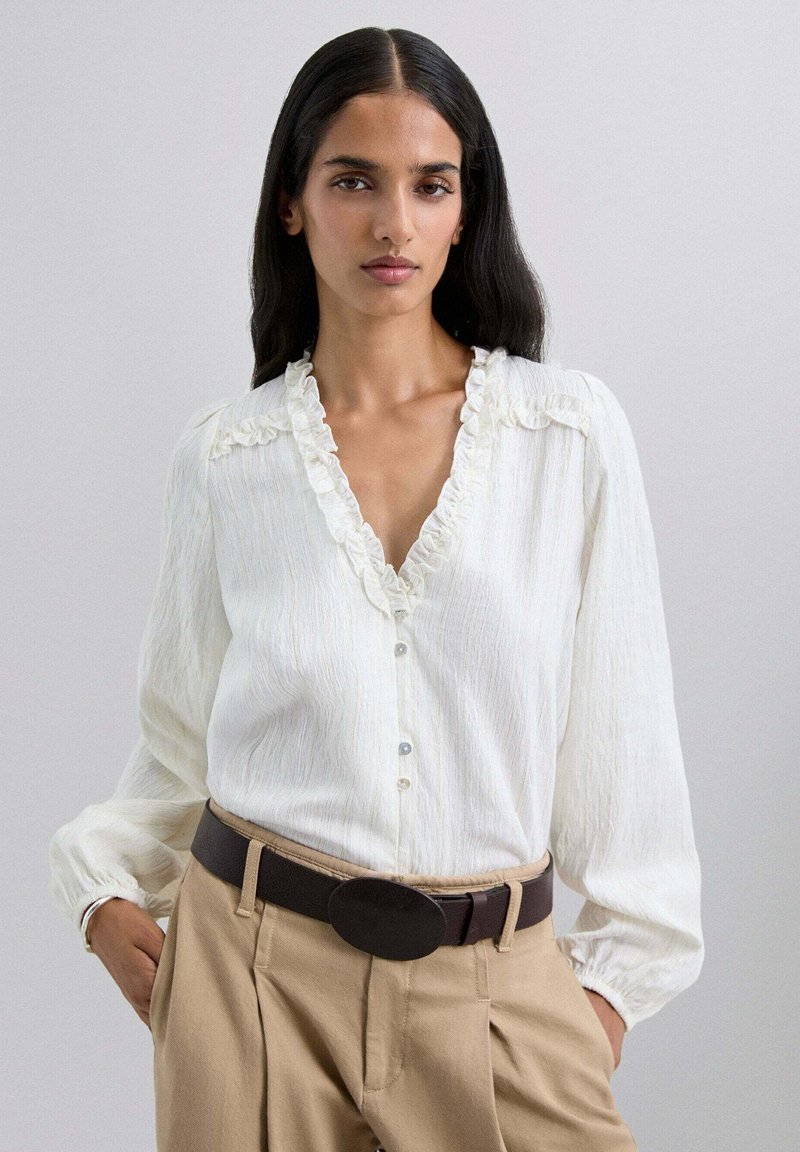 Blusa blanca con un escote en V con volantes, mangas largas abullonadas y cierre de botones, combinada con pantalones beige de cintura alta y un cinturón oscuro.