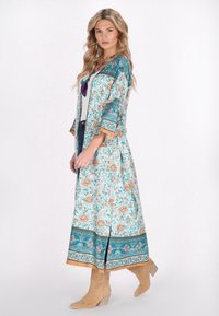 Kimono lungo floreale azzurro chiaro con maniche a tre quarti, dettagli arancioni e orlo con motivo. Indossato su una maglietta beige e denim.