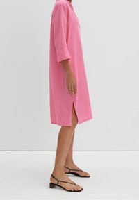 Rosa bomullsklänning med en avslappnad passform, trekvartsärmar, sidoklyftor och knälång längd, i kombination med svarta remsandaler.